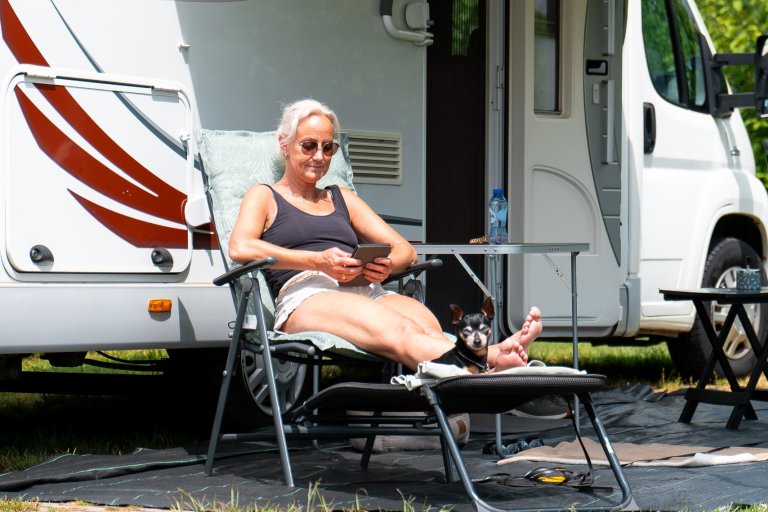 Een vrouw zit maar hondje op een e-reader te lezen voor een camper